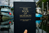 A photo of a copy of The Book of Mormon being held up by a hand against a blurry background on fishing boats in a dock with an overcast blue sky and green hills. The book and hand are in sharp focus and in a different light to the boats, suggesting that the book may be held up to a different photo, or cropped into this one.