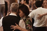 A group of people dance in a room. They are dancing in couples, each couple in a close embrace.