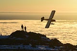 Two people stand on a mountaintop as a single-engine prop plane banks above a sea of clouds…Broken Rainbows by Jim Latham | Photo by Paxson Woelber on Unsplash