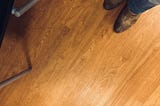 Image of a wood floor with the author’s brown cowboy boots and jeans in the upper right corner, and a part of a chair in the upper left corner.