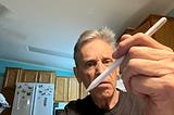 An older man in a kitchen holds an Apple Pencil close to the camera with a curious, slightly skeptical expression on his face. Wooden cabinets and a refrigerator with butterfly magnets are visible in the background.
