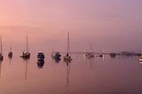 Boats lie at anchor in a peaceful bay. It is evening and the sky is a shade of pinky purple and that is reflected onto the water
