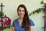 Wearing a blue v-neck blouse with her long, dark hair parted and draped over the front of her left shoulder, Marilys Galindo is pictured in front of a bright, stucco wall with flowering vines trained to provide a colorful but undistracting backdrop to her warm smile.