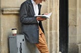 A man standing against a sone wall holding a book in both hands with a coffee at his side.