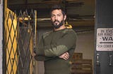 Rudy Reyes with arms crossed in an olive long-sleeved shirt in a warehouse