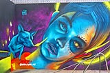 Purple wall with a colorful street art painting — a blue Xoloitzcuintli dog (Aztec dog of the Gods) is on the right shoulder of a blue woman’s reclining head. Her left eye is glowing golden and is surrounded by dark blue coloring and red dots around it.