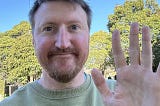 A 40 year old male outside smiling and waving to the camera.