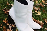 White ankle boots with a three inch heel placed on the grass surrounded by fall leaves.