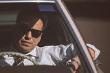 Man wearing formal clothes and sunglasses driving a car.