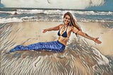 Woman at the beach in a blue bikini top and a blue fabric mermaid tail. Her is long and worn down and she’s wearing a shell necklace. She’s drawn to being a mermaid because doing it says something about myths, mermaids, and women’s power.