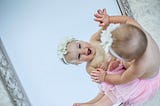 A baby dressed in a pink tutu and a flowery head-band delighting in her reflection in the mirror.