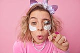 Young woman with messy blonde hair wearing cat ears while curling her eyelashes. Her eyebrows are hi-lo, she’s wearing eye bag patches and a pink sweater against a pink background.