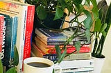 A picture of a white cup of black coffee resting on a bookshelf with books, a money plant, and a snake plant and a red love-shaped deco visible in the background.
