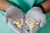 Medical professional wearing green scrubs and light blue latex gloves holding out two handfuls of a variety of pills.