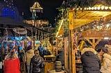 Outdoor Christmas market beer garden at night, with wooden huts, warm fairy lights, people sitting at barrel tables, and a tall lit-up tree in the background.
