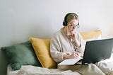 Photo of Woman wearing classes is Sitting on a Couch or Bed and Writing in her Notebook while her Laptop is open