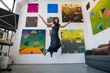 A woman in a long black dress jumps into the air in front of a wall on which 8 colorful geometric abstract paintings are hung.