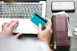 Photo of a person holding a credit card in one hand, while clicking on their laptop with the other one. There is also a wallet visible and a mobile phone.