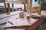 A notebook, a coffee mug and a little milk jug on a wooden table in a coffee shop.