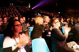 An audience of Interaction Awards attendees applaud. They are seated in a theatre and looking at the stage which is out of frame to the right.