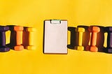 A notepad and with three colorful dumbbells on each side on a yellow background.