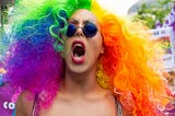 Close up of a woman’s face. She has rainbow hair, sun glasses, and is yelling