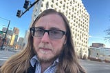 The author has long hair and a beard. He’s wearing sunglasses and a stripped dress shirt with a coat. He’s standing on a street corner in front of a white building with skyscrapers and a streetlight in the background. There are no clouds in the blue sky.