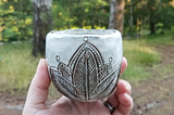 A white and brown,  hand formed ceramic tumbler with an etched mandala-like design, held up against a forested backdrop