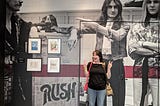 This shows a woman standing in front of a wall with the band Rush painted on it.