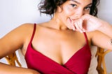 Lovely young woman in wine colored spaghetti-sleeved dress lounging in chair, head titled coquettishly back of right hand held against her mouth. She smiles slightly, seductively, in a come-hitherish way.