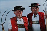 Two old and angry-looking Tyrolean farmers with red vests and pipes in their mouths, holding pitchforks.
