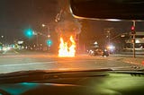 Road intersection with big flames in front of a Mini Cooper with debris scattered on the floor.