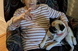 An elderly woman in a striped shirt and dark pants sits in an arm chair with a pattern in shades of green. She is pointing with her right hand and holding a young pug on her left side.