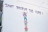 “Don’t believe the hype” graffiti on a white brick wall