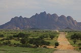 A gravel road leading between grassy meadows toward a granite mountain in the background.