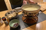 A pot of cheese sits on a burner on the table along with chopped bread and pieces of cured meats.