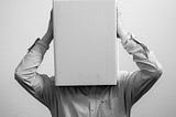 A black and white image of a man in a shirt holding a box that he has placed over his face and head