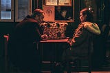 Photo Description: An older couple sit together in a dark diner having a discussion. They both look unhappy and still have their coats on. No food is on the table.
