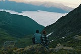 Photo of a man and woman together on a dark, high vista, looking at a lighted valley in the mountains with a lake.