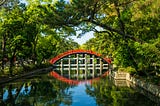 A photo showing green tree foliage over an arched red walking bridge reflected in the still water