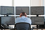 A frustrated worker with their hands on their head sits down at their desk, which has six computer monitors.