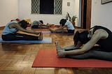 Pessoas praticando yoga sentadas no chão na posição da pinça, com pernas juntas estendidas e a coluna flexionada com as mãos na direção dos pés.