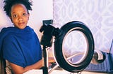 Woman sitting in front of her laptop. She is next to a camera light. She looking towards the camera and smiling.