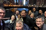 A photograph of eight men and five women, smiling, wearing winter clothing, standing on a bridge over the Chicago river, taken at dusk. Several large buildings — and another bridge — are visible in the background.
