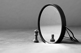 A black and white image of a knight chess piece in front of a small mirror, the reflection shows a pawn.