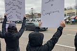 Two women dressed in black raincoats hold up white signs, one with “Silence = Compliance” and the other filled with a litany of issues (including but not limited to “Social Security,” “Education,” “Health Care,” “Research,” “History,” and “National Parks”), for passing cars on a wide avenue to see. Across the way, are other demonstrators with colorful handmade signs, including “RESIST.” (Photo by author.)