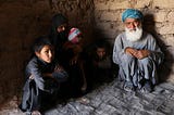 an old man, a woman in a hijab and three small children huddle in a corner of a brick house