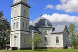 Colour photo from 2014 of Rautjärvi Lutheran church, Finland. A wooden church with a square wooden church tower topped by a metal-roofed spire. The church is painted light grey with white trim.