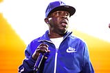Rapper Phife Dawg holding a microphone on stage, wearing a blue baseball cap and matching track jacket