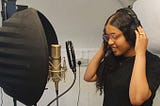 Malizah, standing next to recording kit, she is a oung Black woman with long black haid, and glasses. She is holding her headphones at each ear.